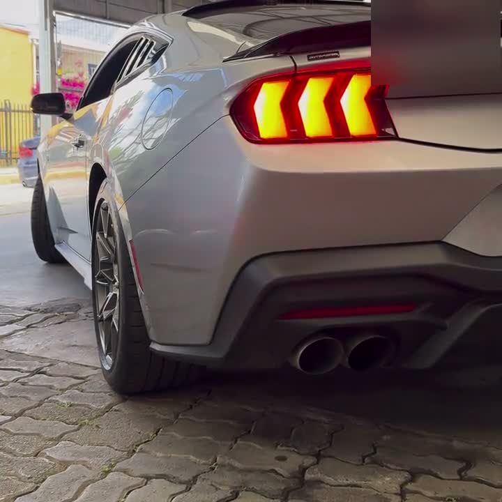 Parte trasera de un Ford Mustang gris con las luces encendidas, en el patio de una automotora de la red.